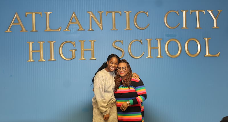 Two women pose in front of a blue wall with "ATLANTIC CITY HIGH SCHOOL" written in gold lettering.