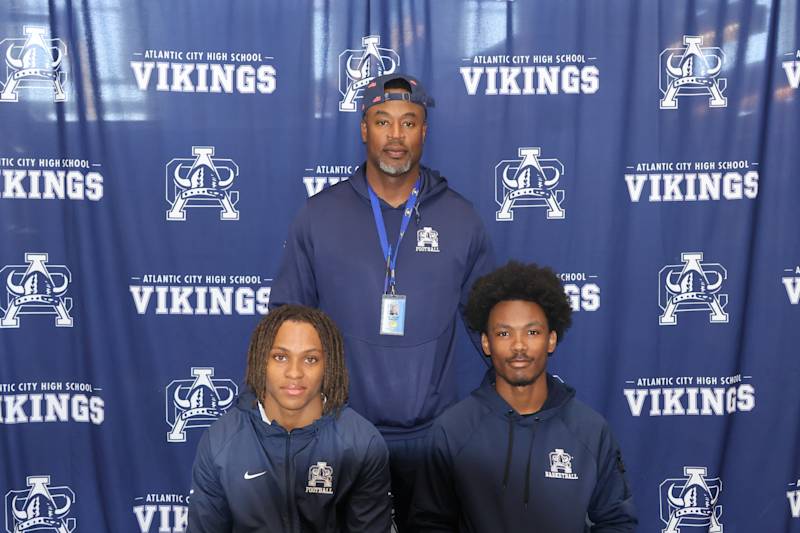 A man stands behind two young men, all posing in front of a blue backdrop with "Atlantic City High S…