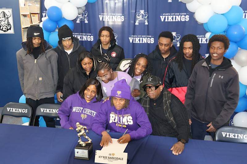 A group of people, some wearing purple, gather around a table with a signing document in the foregro…