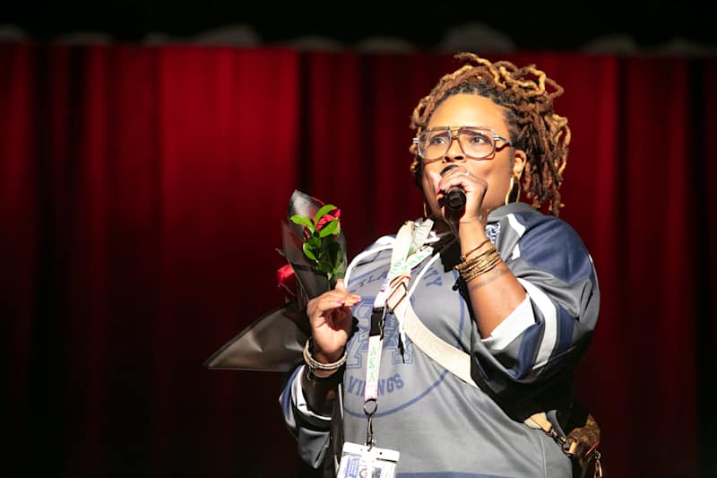 A woman with dreadlocks and glasses holds a bouquet of red roses while singing into a microphone on…