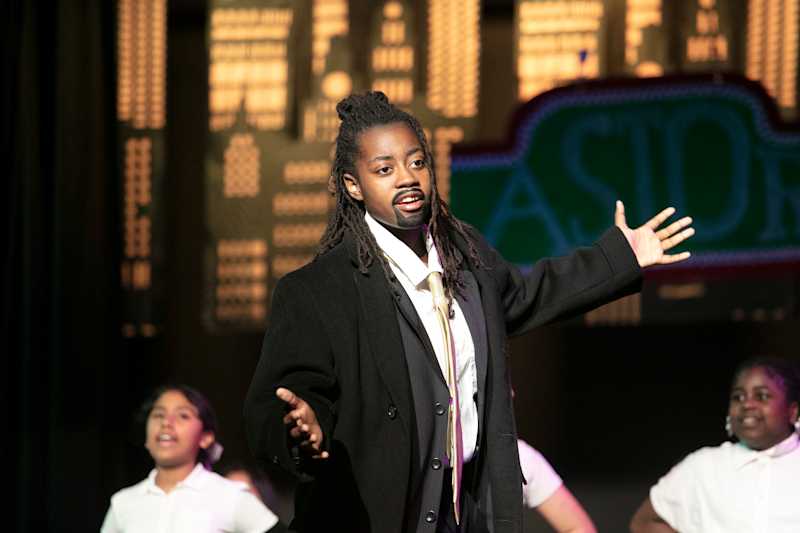 A young person in a costume with a fake beard and a suit stands center stage with arms outstretched,…