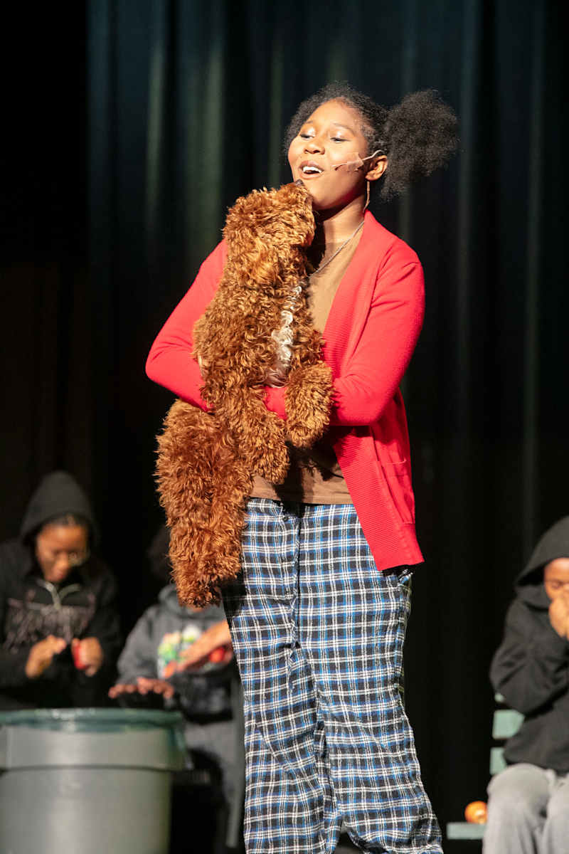 A young woman in plaid pajamas and a red cardigan sings while holding a stuffed dog on a stage with…