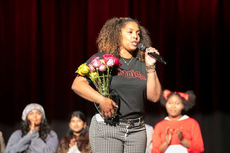 A woman with a bouquet of roses speaks into a microphone on a stage with a red curtain backdrop and…
