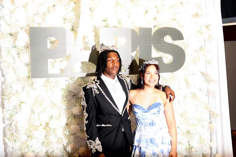 A young couple in formal attire poses in front of a white floral wall with large silver letters spel…