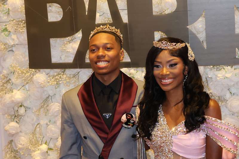 A smiling young couple, adorned with crowns, poses in front of a white floral wall and a large "PAR…