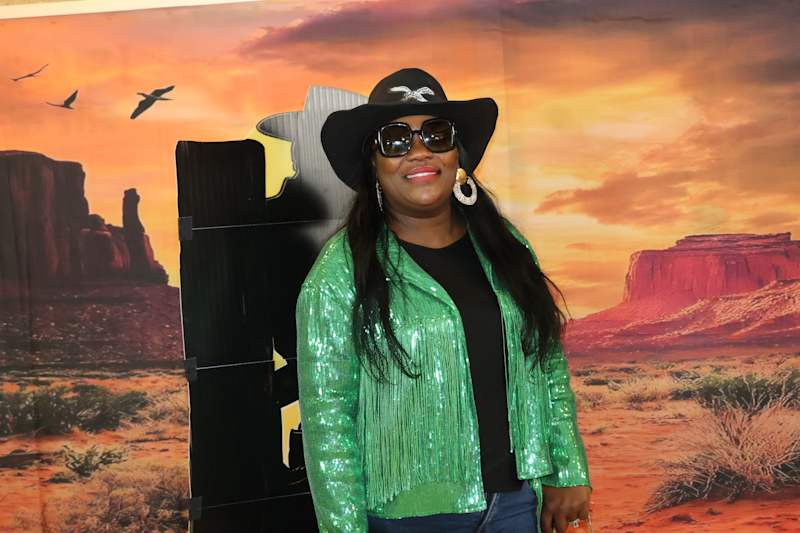 A woman in a sparkly green jacket and cowboy hat poses in front of a desert landscape backdrop.