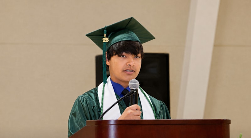 A young man in a graduation cap and gown speaks at a podium in front of a plain wall.