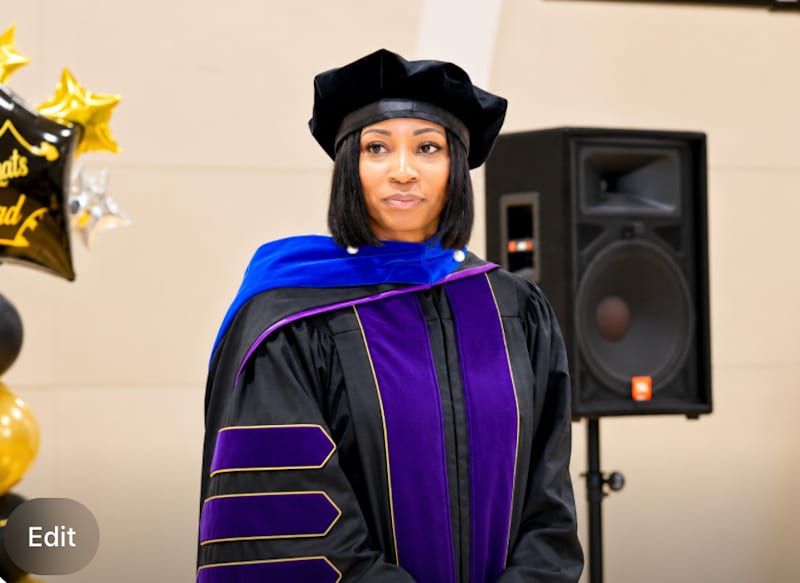 A woman in academic regalia stands in the foreground with a speaker and balloons in the background.