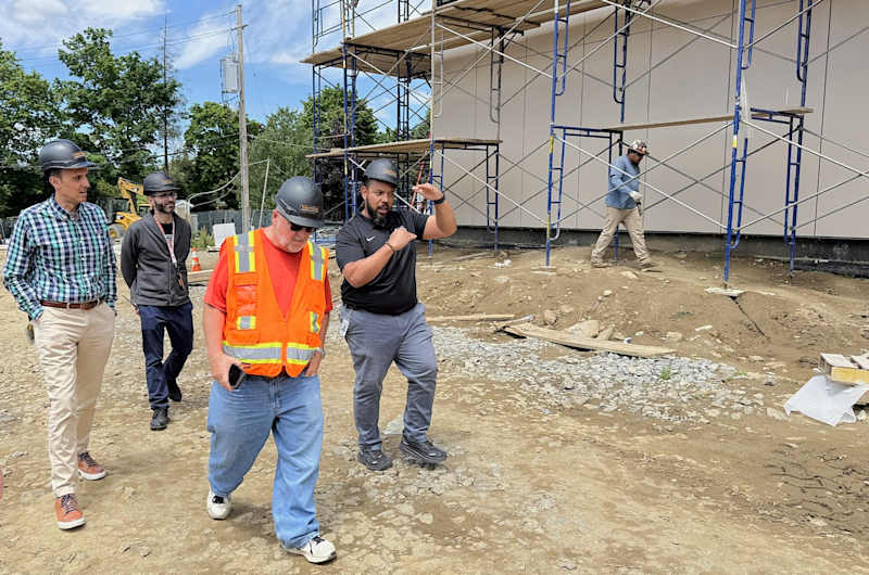 Four men in hard hats walk on a gravel path in the foreground, while a building under construction with scaffolding stands in the background.