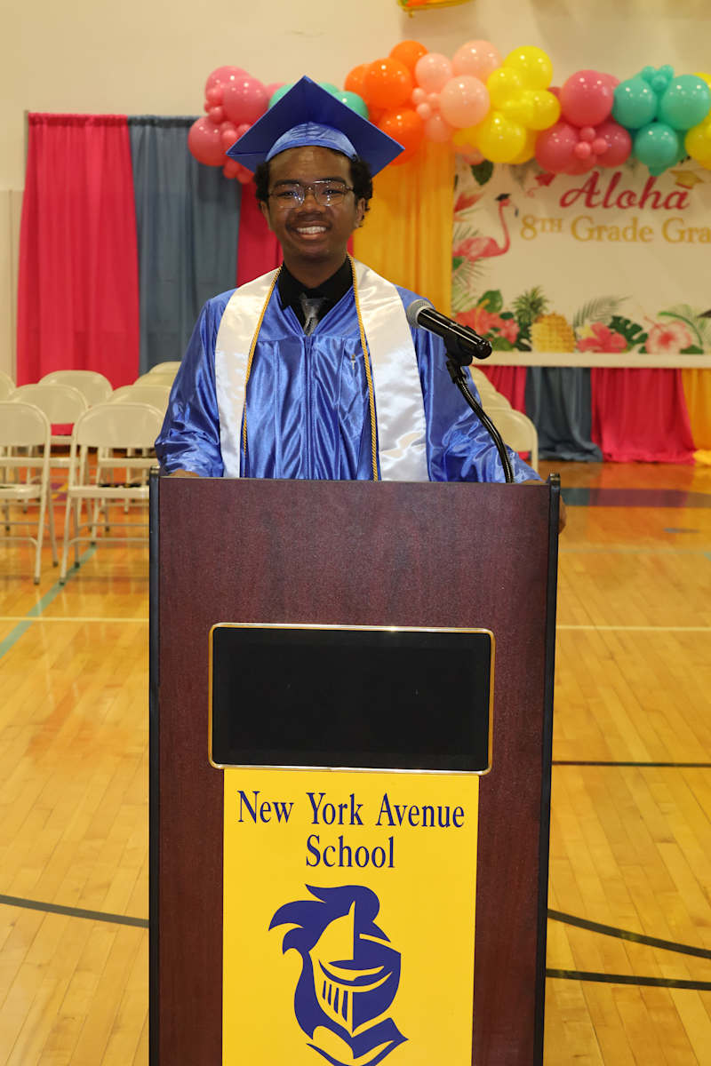 A smiling graduate in a blue cap and gown stands at a podium with the "New York Avenue School" logo,…
