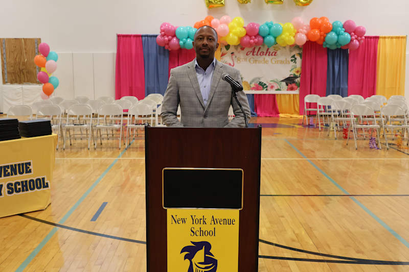 A man stands at a podium in front of a colorful balloon arch and rows of chairs, with a yellow banne…