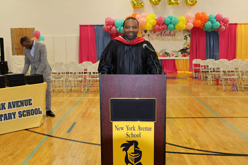 A man in a graduation gown stands at a podium in front of a decorated backdrop with balloons and ban…
