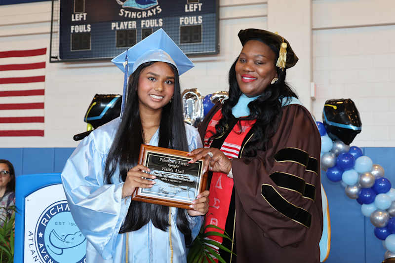 A young woman in a light blue graduation gown and cap receives a plaque from a woman in academic reg…