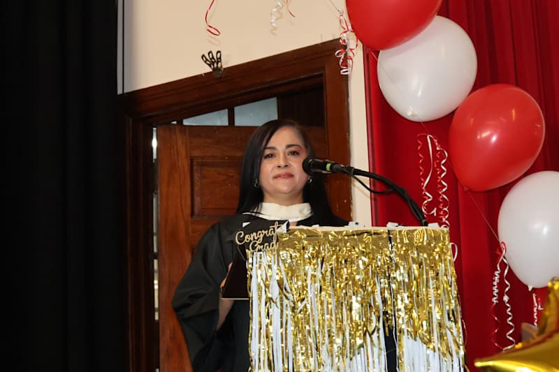 A woman in a graduation gown stands at a podium decorated with gold fringe, with red and white ballo…