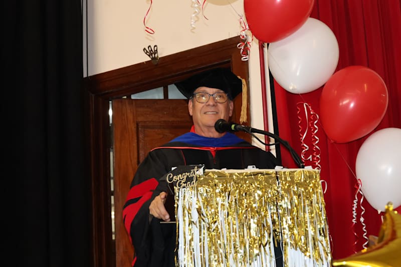 A man in academic regalia stands at a podium decorated with gold tinsel and balloons, with a doorway…
