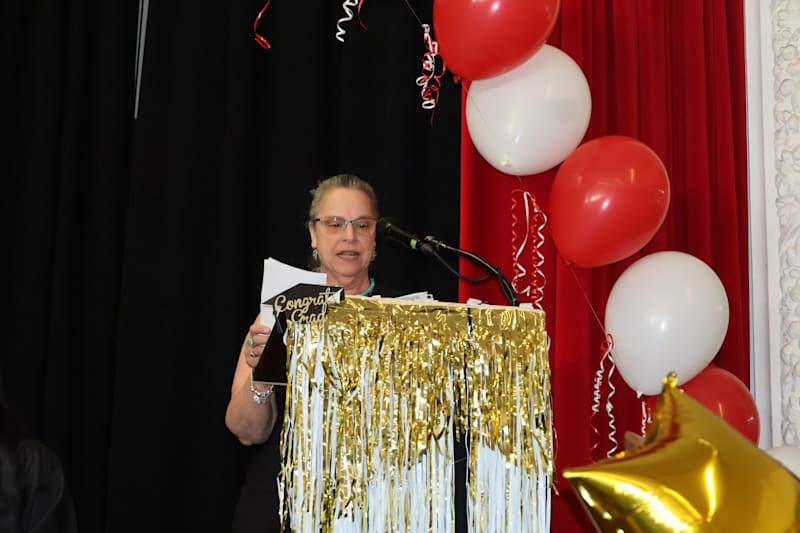 A woman speaks at a podium adorned with gold fringe, while red and white balloons float against a re…