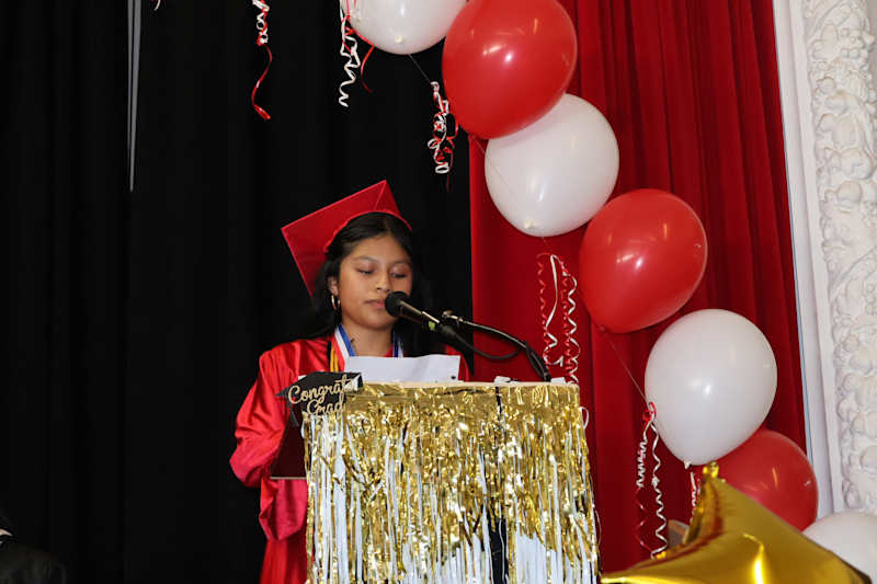A young girl in a red graduation gown and cap speaks at a podium adorned with gold fringe, while red…