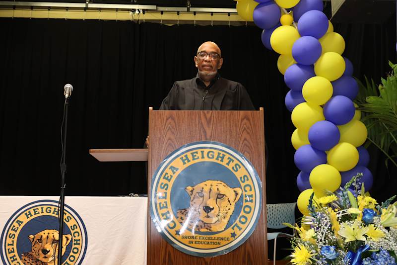 A man stands at a podium with the Chelsea Heights School logo, while a balloon arch and floral arran…