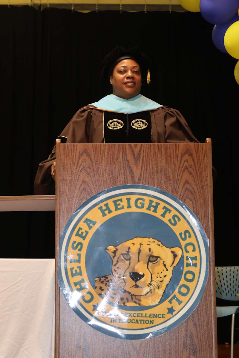 A woman in academic regalia stands behind a podium with a school logo, while balloons are visible in…