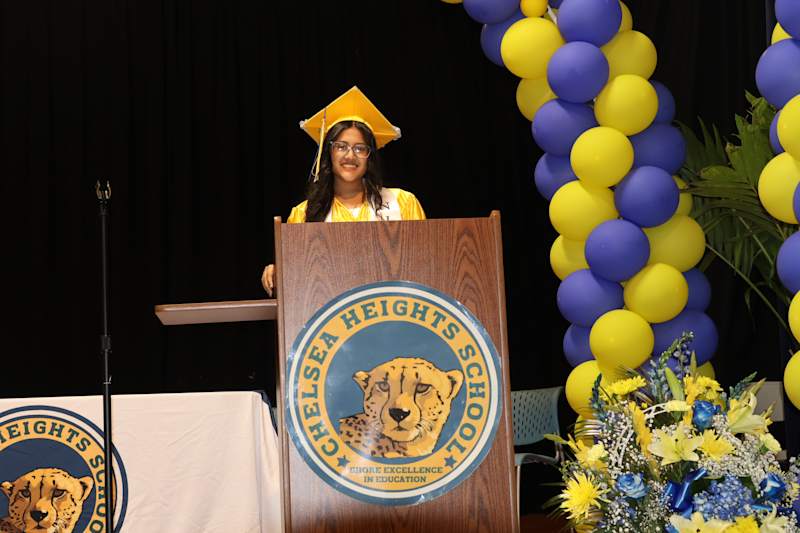 A young woman in a yellow graduation cap and gown stands at a podium with the Chelsea Heights School…