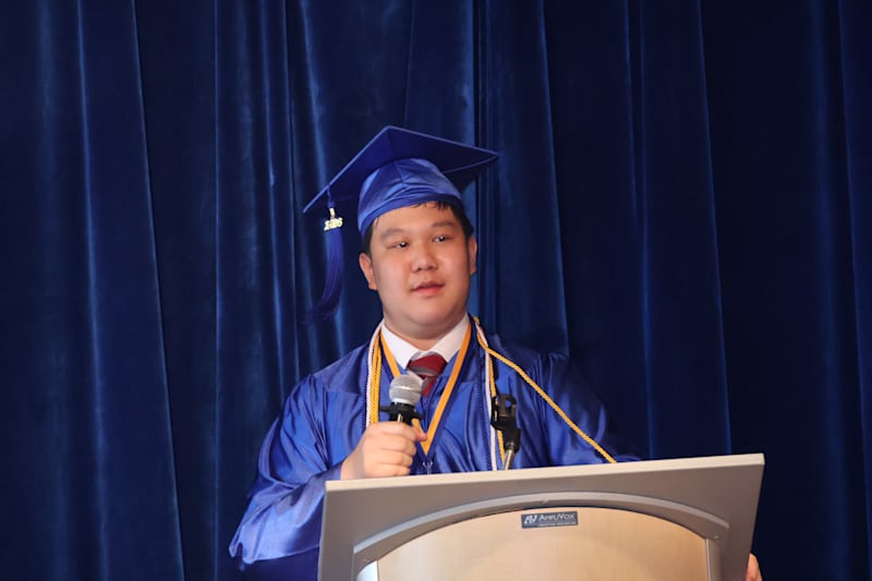 A young man in a blue graduation gown and cap stands at a podium speaking into a microphone, with a…