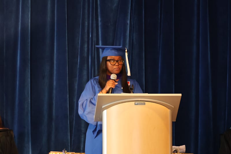 A graduate in a blue cap and gown speaks at a podium in front of a blue curtain.