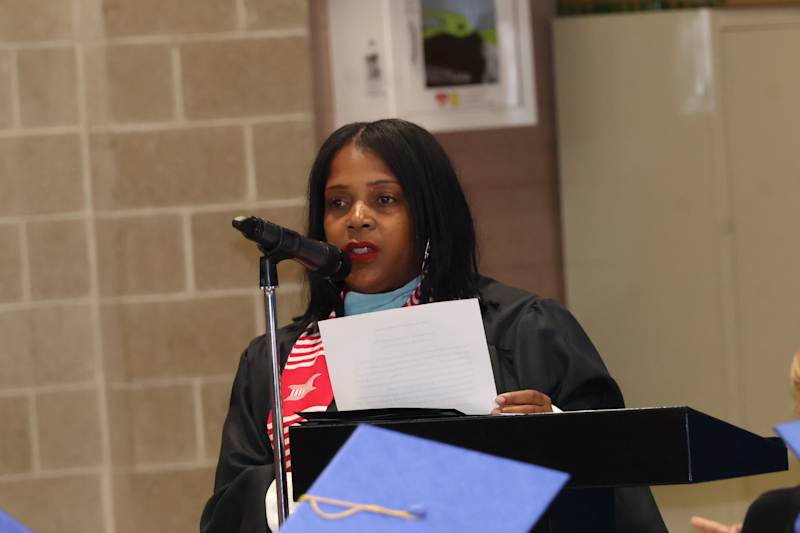 A woman in academic regalia speaks into a microphone at a podium, with a brick wall and a bulletin b…