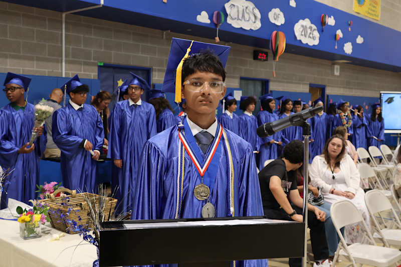 A young man in a graduation cap and gown stands at a podium in the foreground, while other graduates…