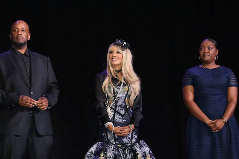 Three people stand on a dark stage, with a man in a suit on the left, a woman in an elaborate dress…