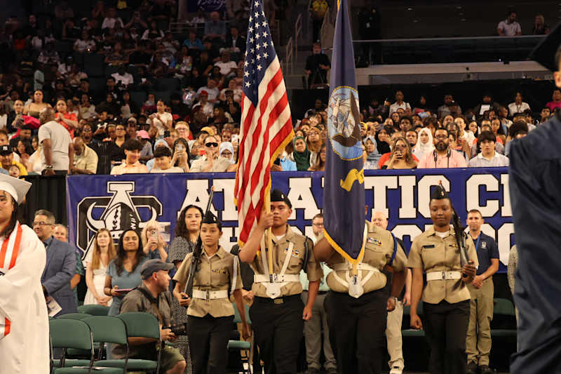 A color guard with American and Navy flags marches in the foreground before a crowd of spectators at…