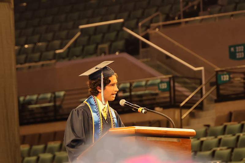 A young man in a graduation cap and gown speaks at a podium in front of an audience seated in a stad…