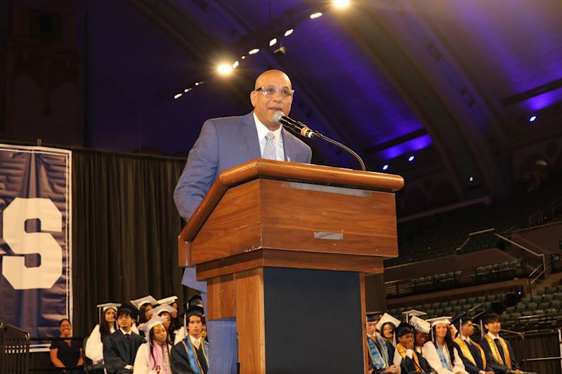 A man in a suit speaks at a podium in the foreground, while graduates in caps and gowns sit in the b…