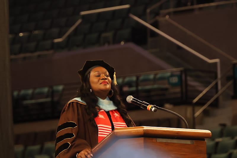 A woman in academic regalia stands at a podium with her eyes closed, while an empty auditorium with…