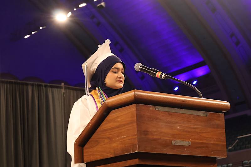 A young woman in a graduation cap and gown speaks at a wooden podium in front of a dimly lit auditor…