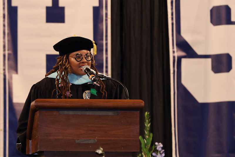 A woman in academic regalia speaks at a podium in front of a large banner.