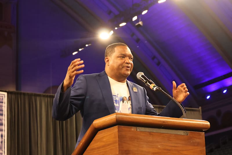 A man in a blue suit speaks at a wooden podium with a purple-lit background.