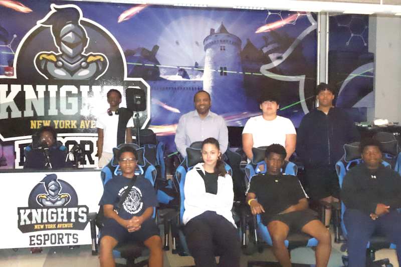 A group of young people and an adult pose in front of a backdrop featuring a castle and gaming logos…