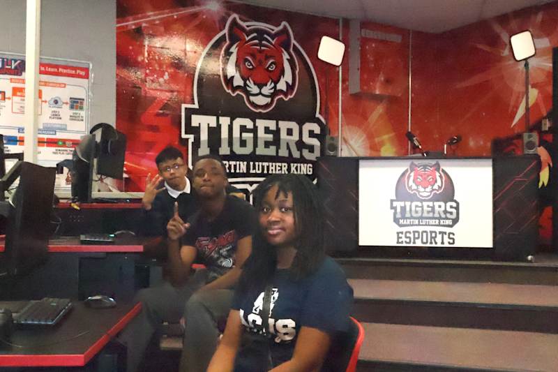 Three young people are seated in front of a large "Tigers Martin Luther King Esports" logo, with gam…