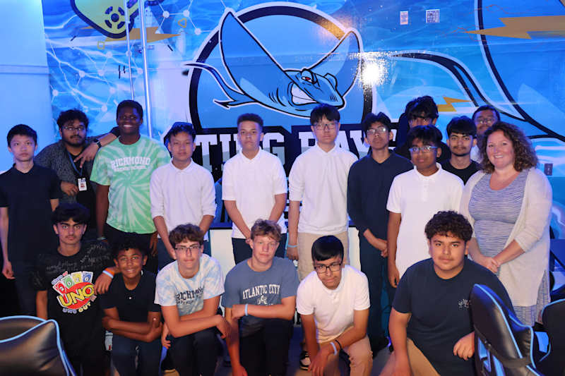 A group of young people and an adult woman pose for a photo in front of a large blue stingray logo.