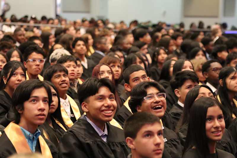 A diverse group of young people in choir robes sing in front of a blurred background with orange accents.