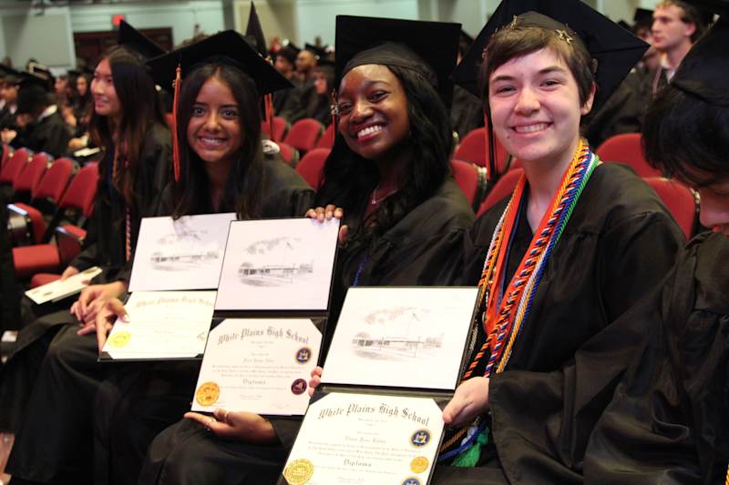 Three smiling graduates in black caps and gowns hold their diplomas in the foreground, while a blurred audience fills the red seats in the background.