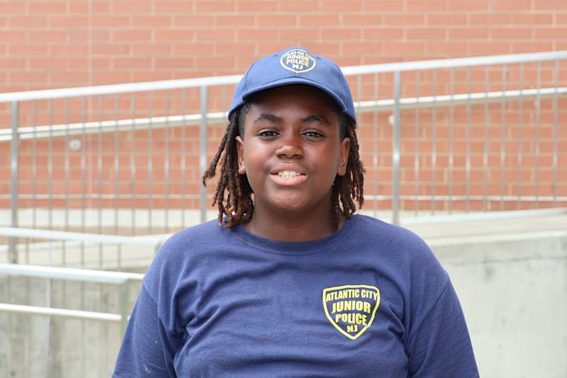 A young person in a junior police uniform stands in front of a brick building with metal railings.