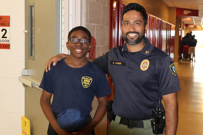 A young boy in a blue t-shirt stands with his arm around a police officer in uniform, with lockers a…