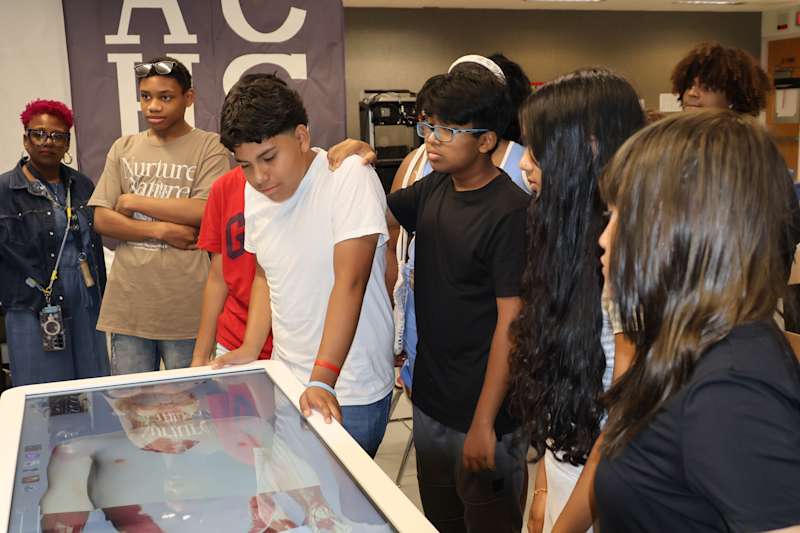A group of diverse teenagers gather around a large interactive screen displaying anatomical models,…