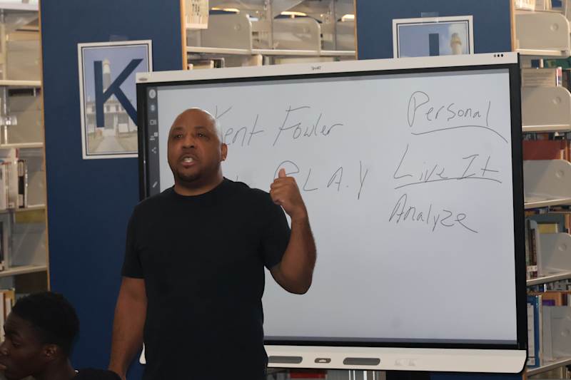 A man in a black t-shirt gestures towards a whiteboard with writing on it, while bookshelves filled…