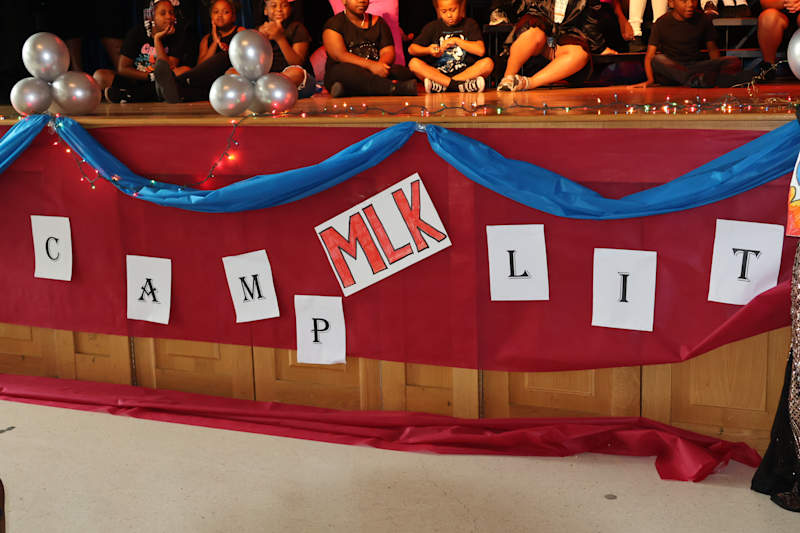 A stage decorated with red and blue fabric, balloons, and letters spelling "CAMP MLK LIT" is in the…