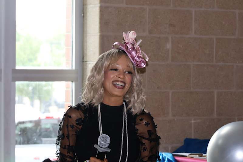 A smiling person in a black outfit and a pink fascinator holds a microphone in the foreground, with…