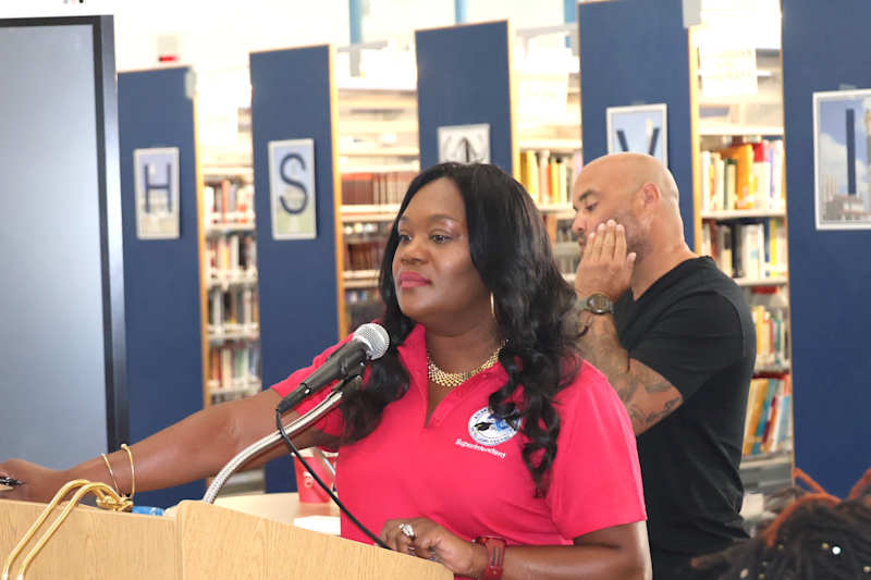 A woman in a pink shirt speaks at a podium in the foreground, while a man with tattoos stands behind…