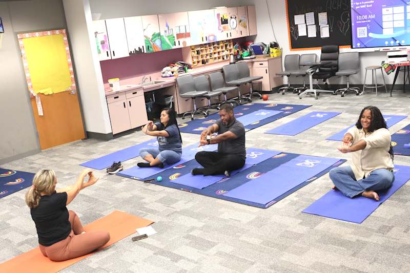 Four people practice yoga on mats in a brightly lit room with colorful decorations on the walls and…