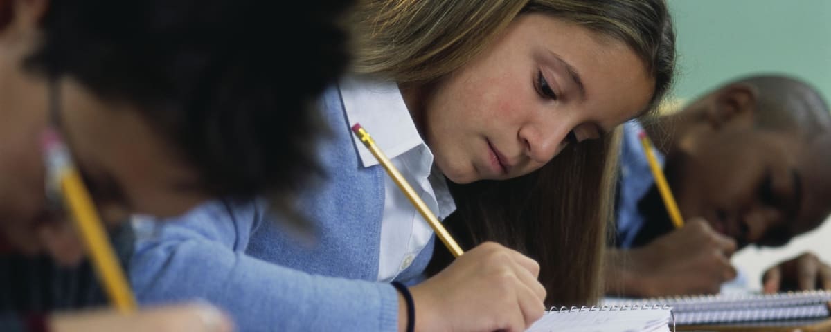 A young woman with long blonde hair is intently focused on writing in a notebook, surrounded by a dark background.